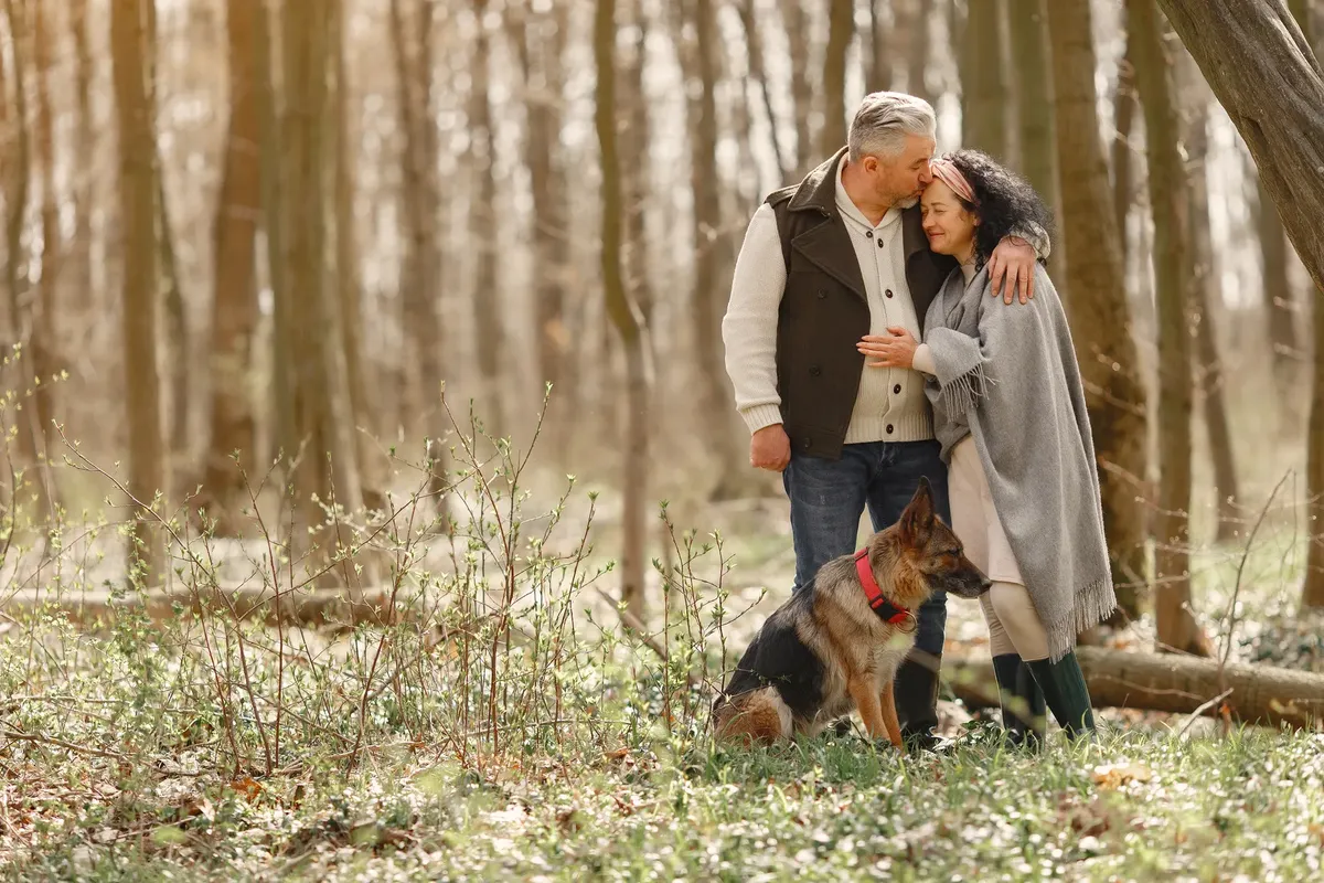 Cheerful senior couple in casual outfit standing together near obedient dog and hugging with tenderness while resting in park in autumn